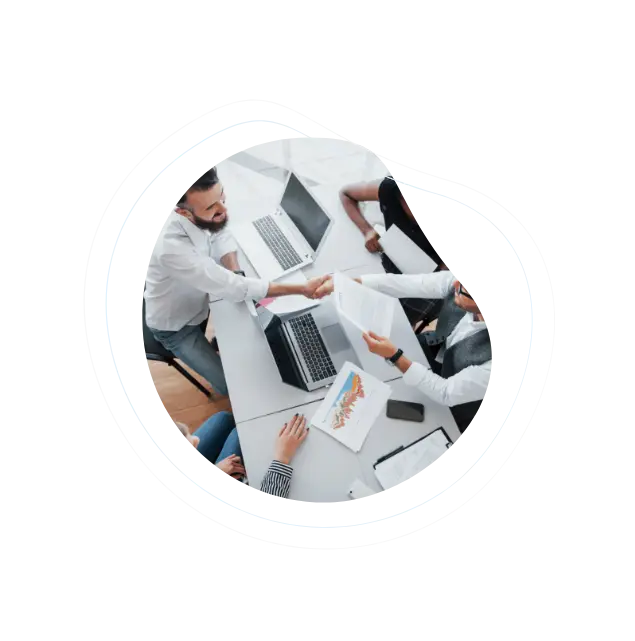 Partnership business professionals shaking hands across a table with laptops and documents, symbolizing collaboration and agreement.